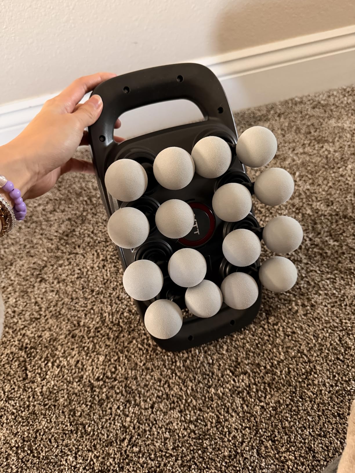 Hand holding a black handheld device with white spherical nodes on a carpeted floor.